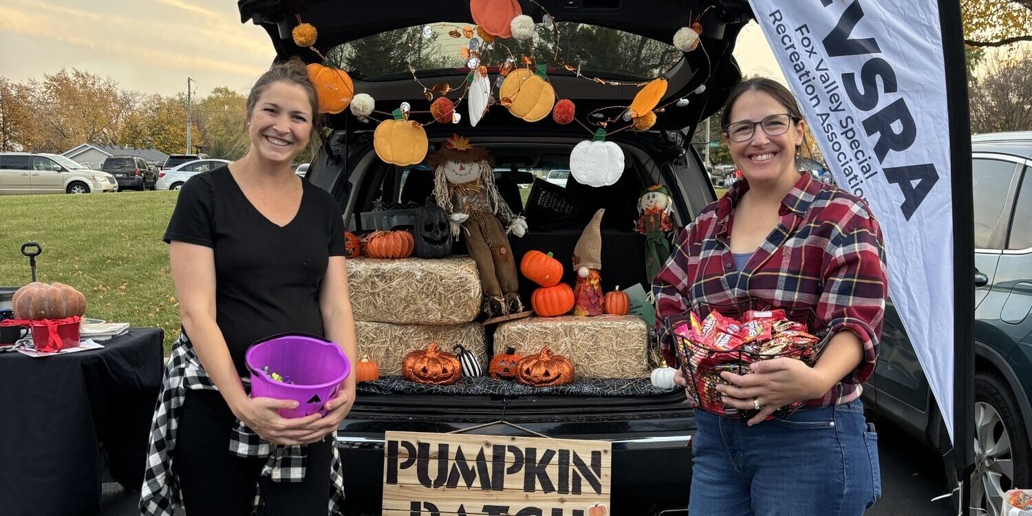 Two women posing with candy buckets next to a fall-themed decorated car trunk at a Trunk-Or-Treat