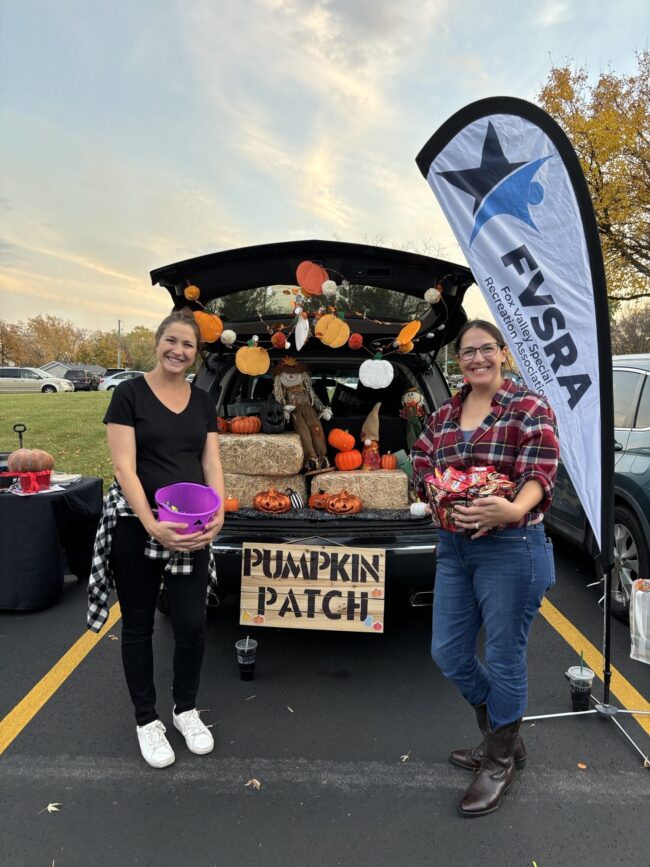 Two women posing with candy buckets next to a fall-themed decorated car trunk at a Trunk-Or-Treat