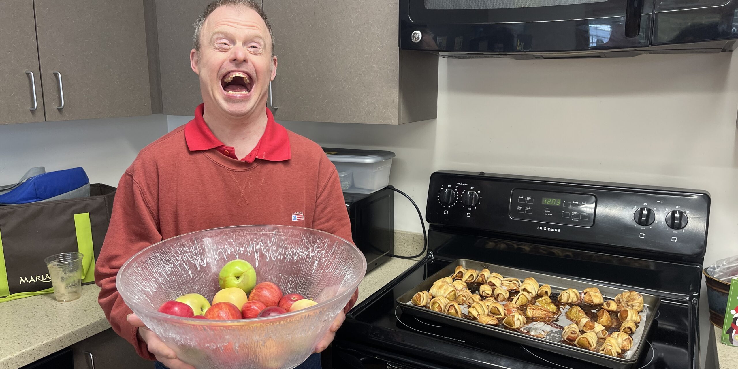 Individual posing by apple pastries on a stovetop while holding a bowl of apples