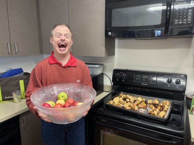 Individual posing by apple pastries on a stovetop while holding a bowl of apples
