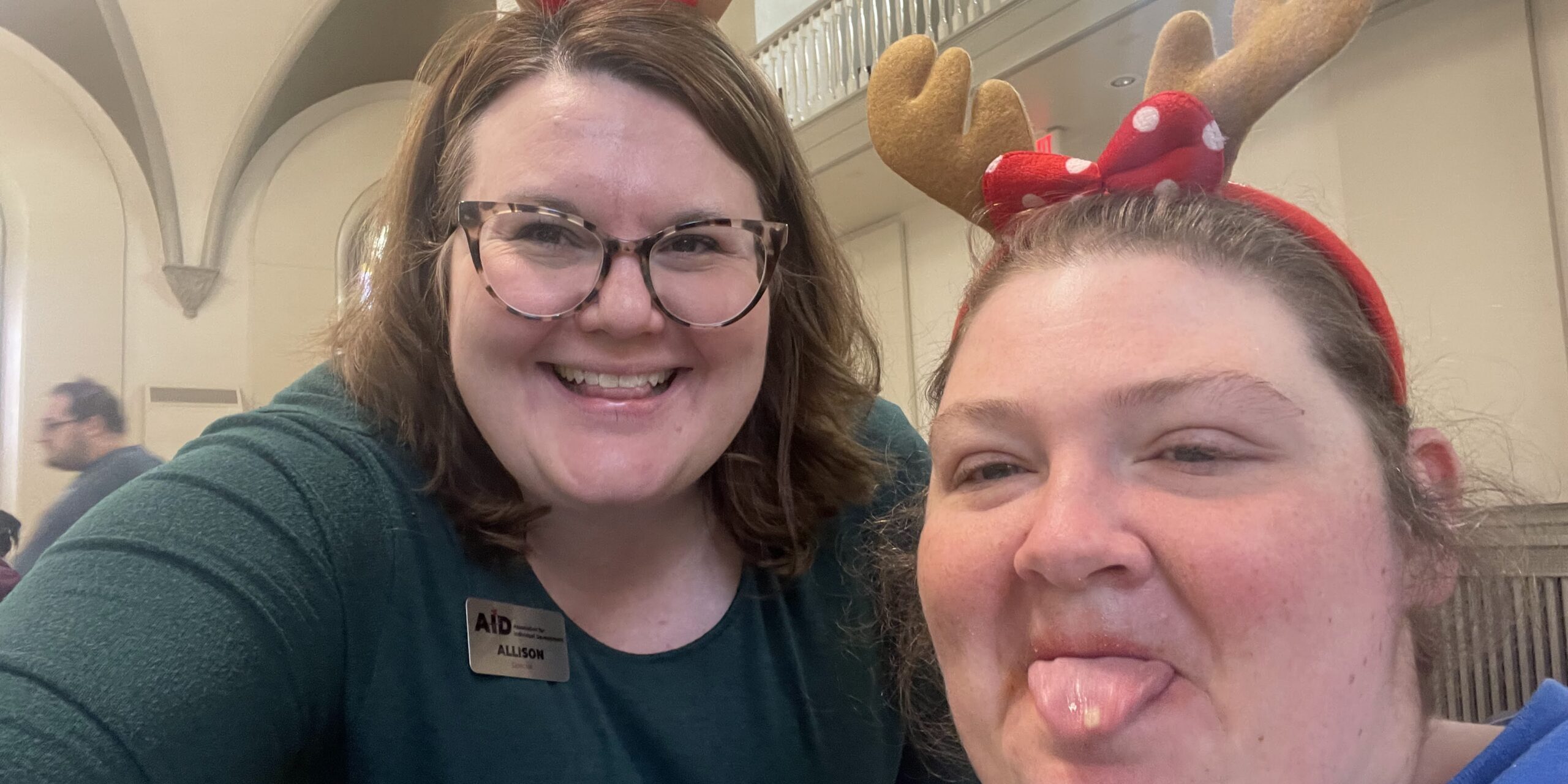 Two women taking a selfie- one smiling and one sticking her tongue out- wearing reindeer antler headbands