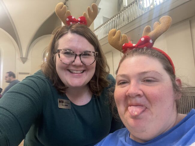 Two women taking a selfie- one smiling and one sticking her tongue out- wearing reindeer antler headbands