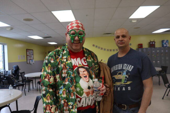 Man decked out in Elf clothing and upside-down Christmas tree glasses poses with another man