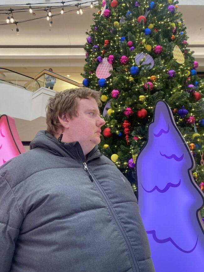 A man waiting in line to see Santa with a very large Christmas tree in the background