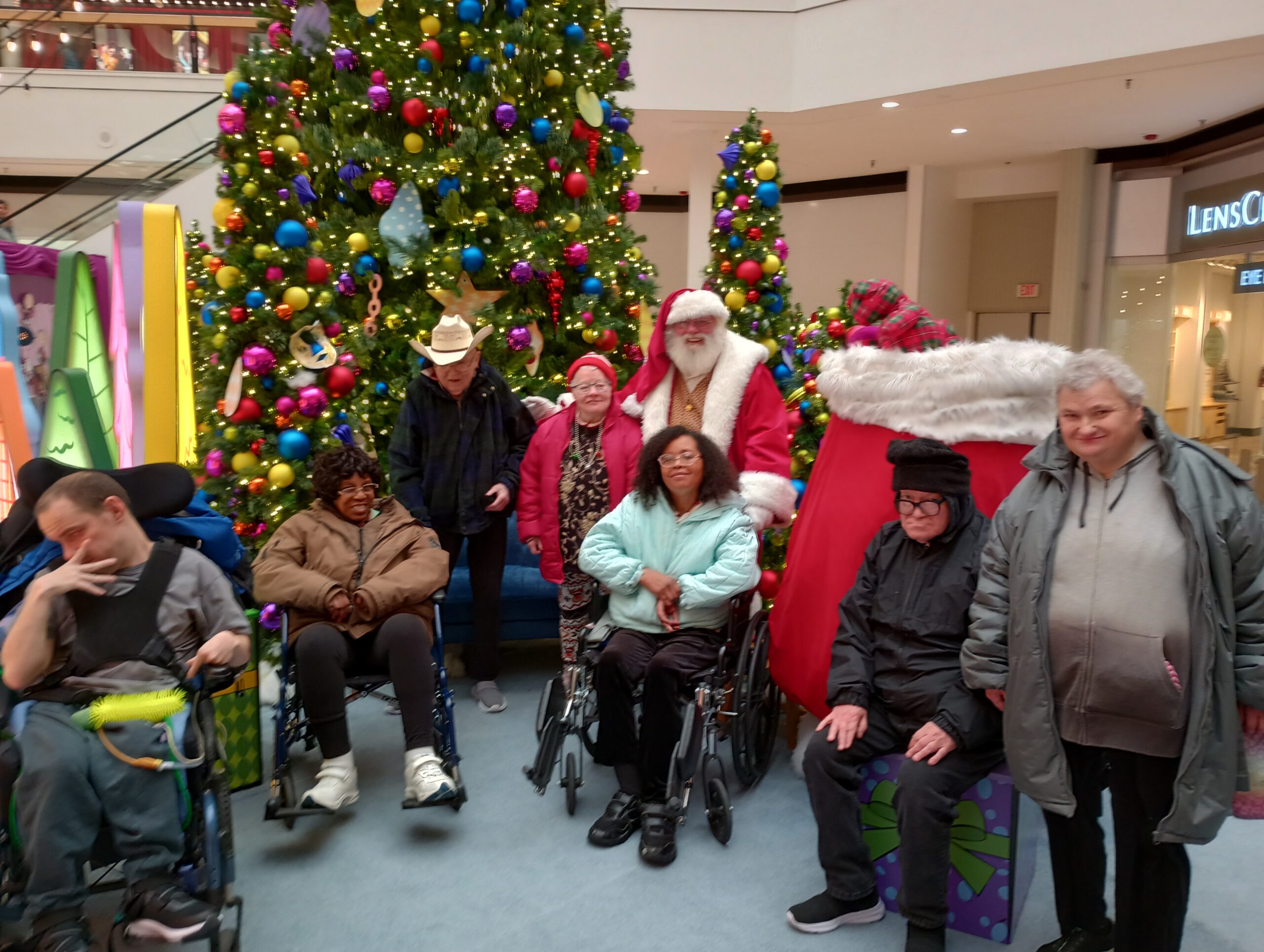 Seven individuals pose with Santa in front of a very large Christmas tree and a smaller Christmas tree at a mall